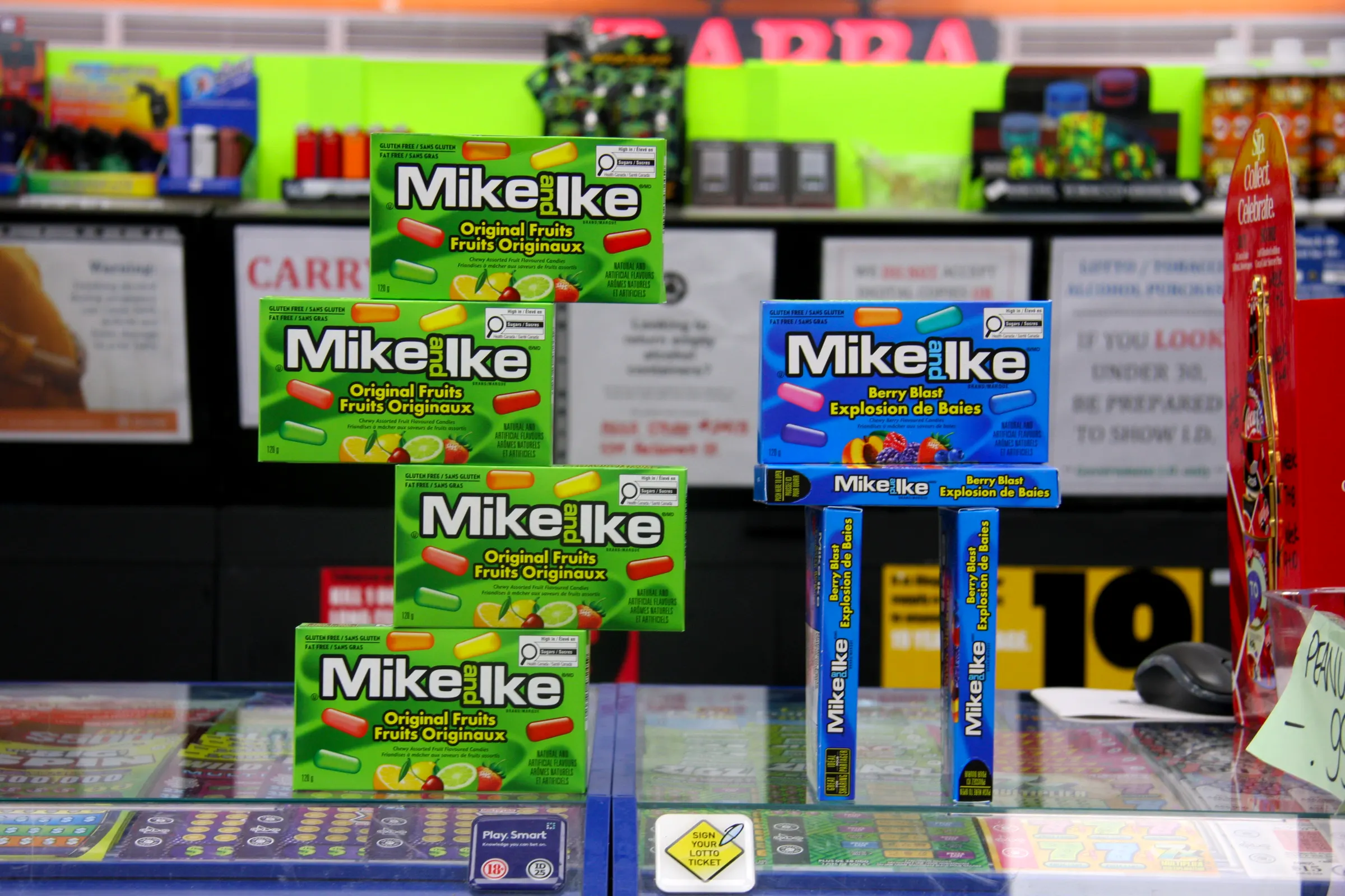 Boxed candy stacked into a tower on the counter glass, lottery tickets laid out beneath it.