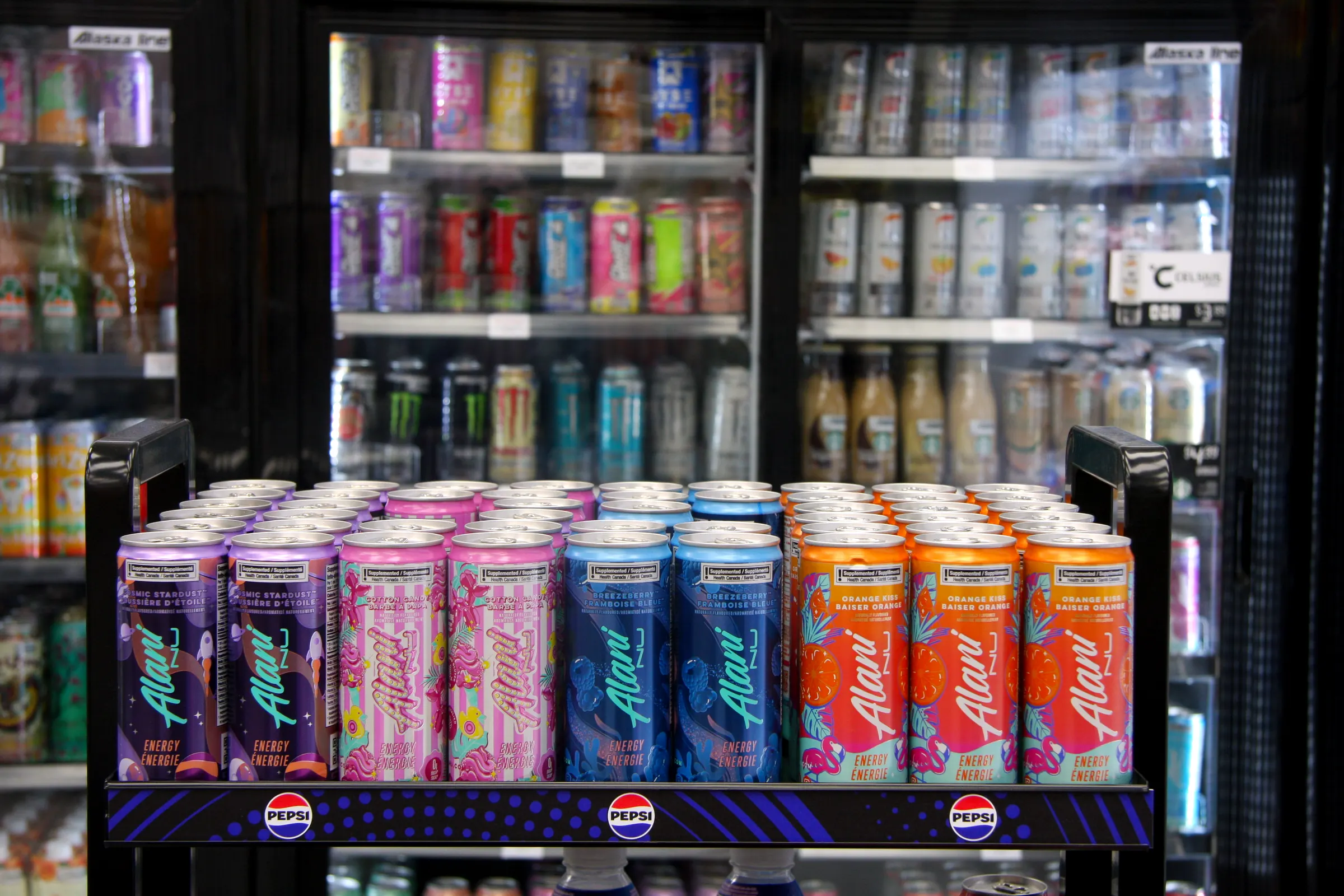 The energy drink display from the side, flavours ranked in colour order against the lit coolers.