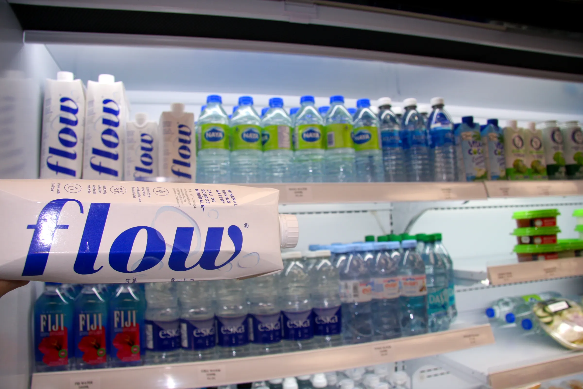 A carton of Flow spring water lifted from the cooler, bottled water ranked on the shelves behind.