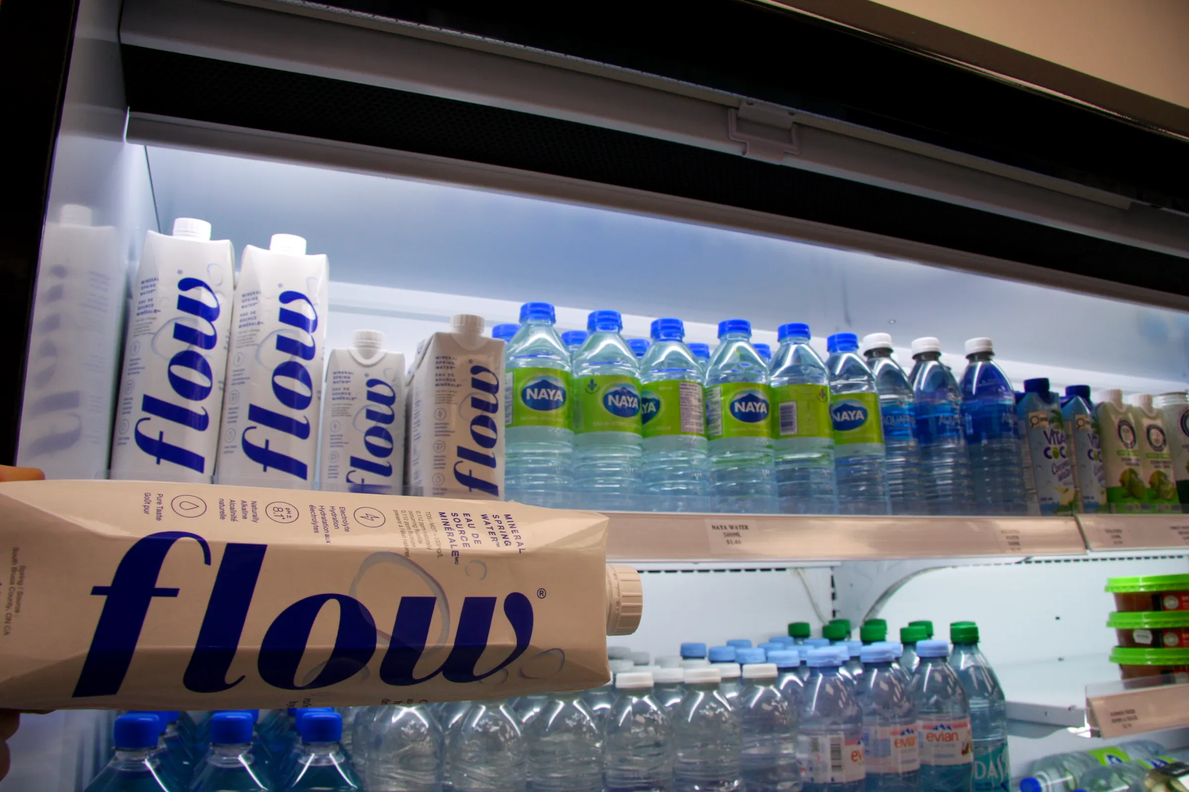 A Flow water carton close in the cooler light, bottled water and juice ranked along the shelf behind.